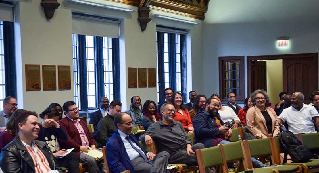 People are seated in a semi-circle in green chairs laughing with smiles on their faces.