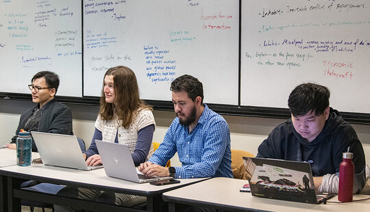 Four people at a table with laptops at the front of the room with a whiteboard behind them.